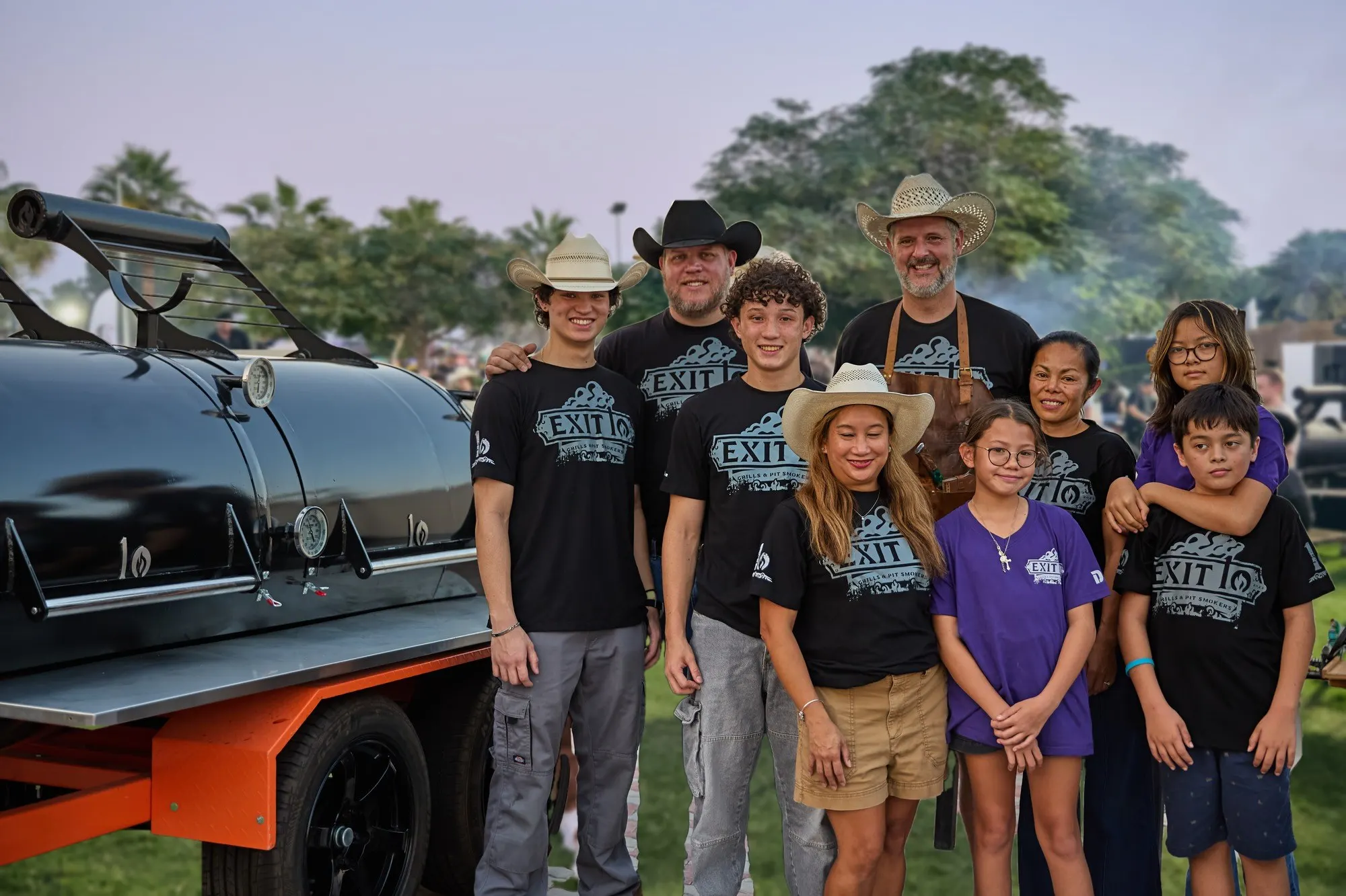 The Exit 10 BBQ family in Exit 10 shirts with Pitmasters Hunter and Dave beside the Smoky Bandit trailer smoker at an Exit 10 BBQ event in the UAE